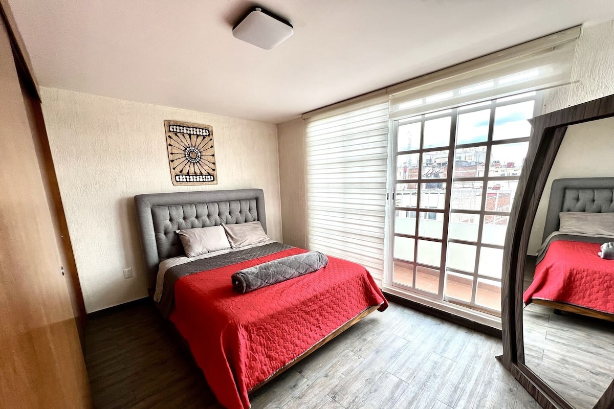Une chambre avec un grand lit, une fenêtre avec des stores, un luminaire au plafond et un élément décoratif mural.