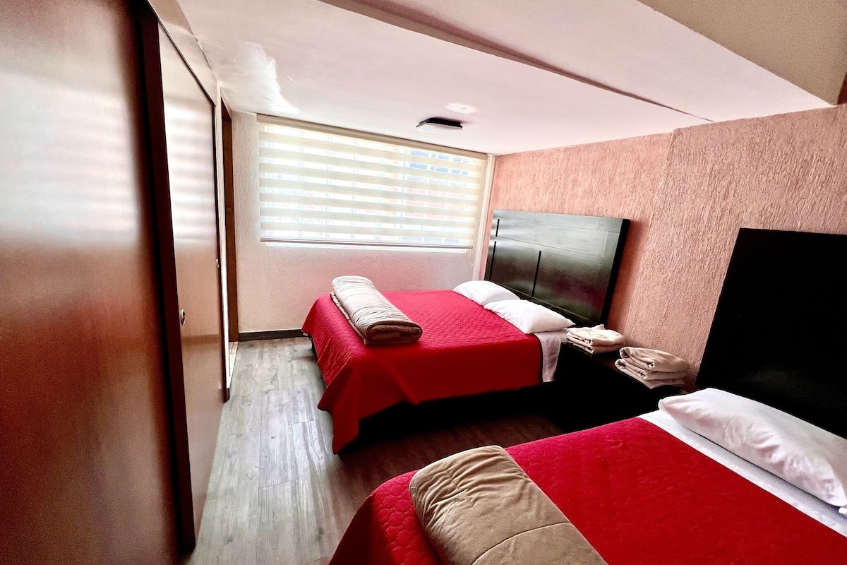 Une chambre d’hôtel avec deux lits, une grande fenêtre avec des stores, un plancher en bois et une tête de lit sombre.