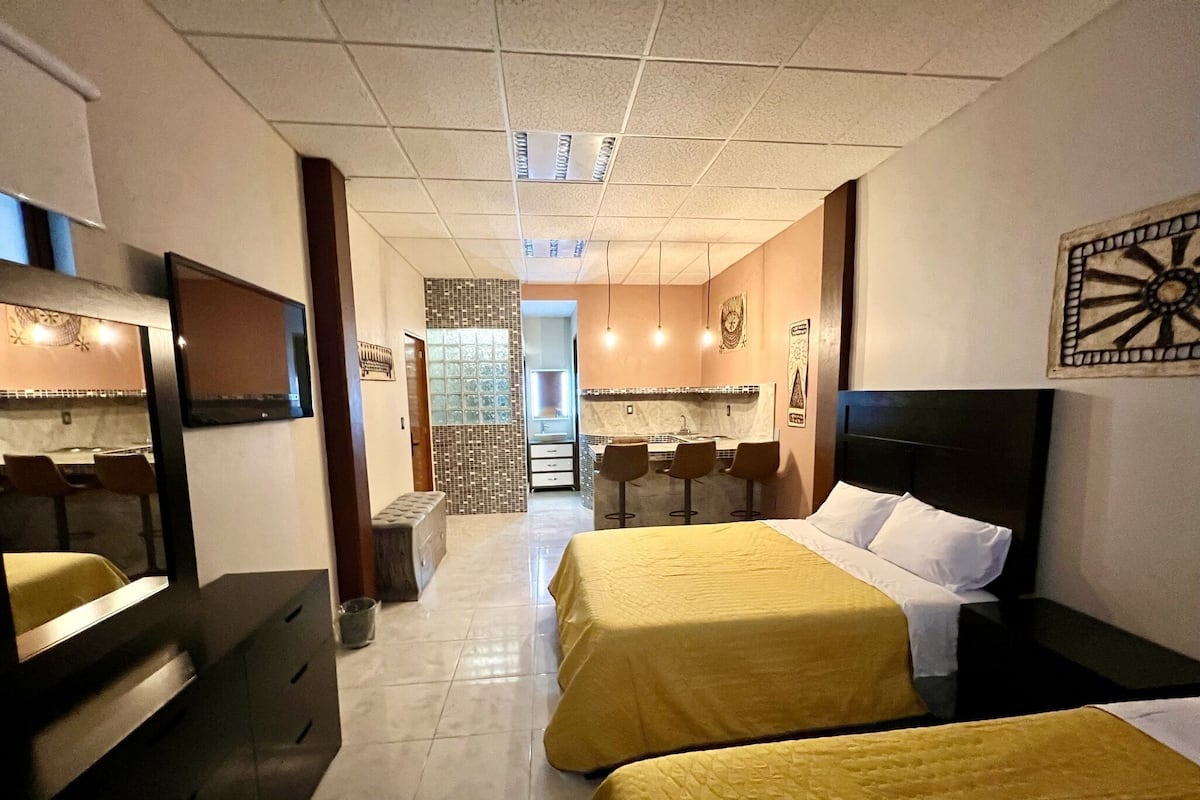 Une chambre d’hôtel avec deux lits, un coin salon avec une table et des chaises, une kitchenette et une salle de bain avec un miroir et un lavabo.