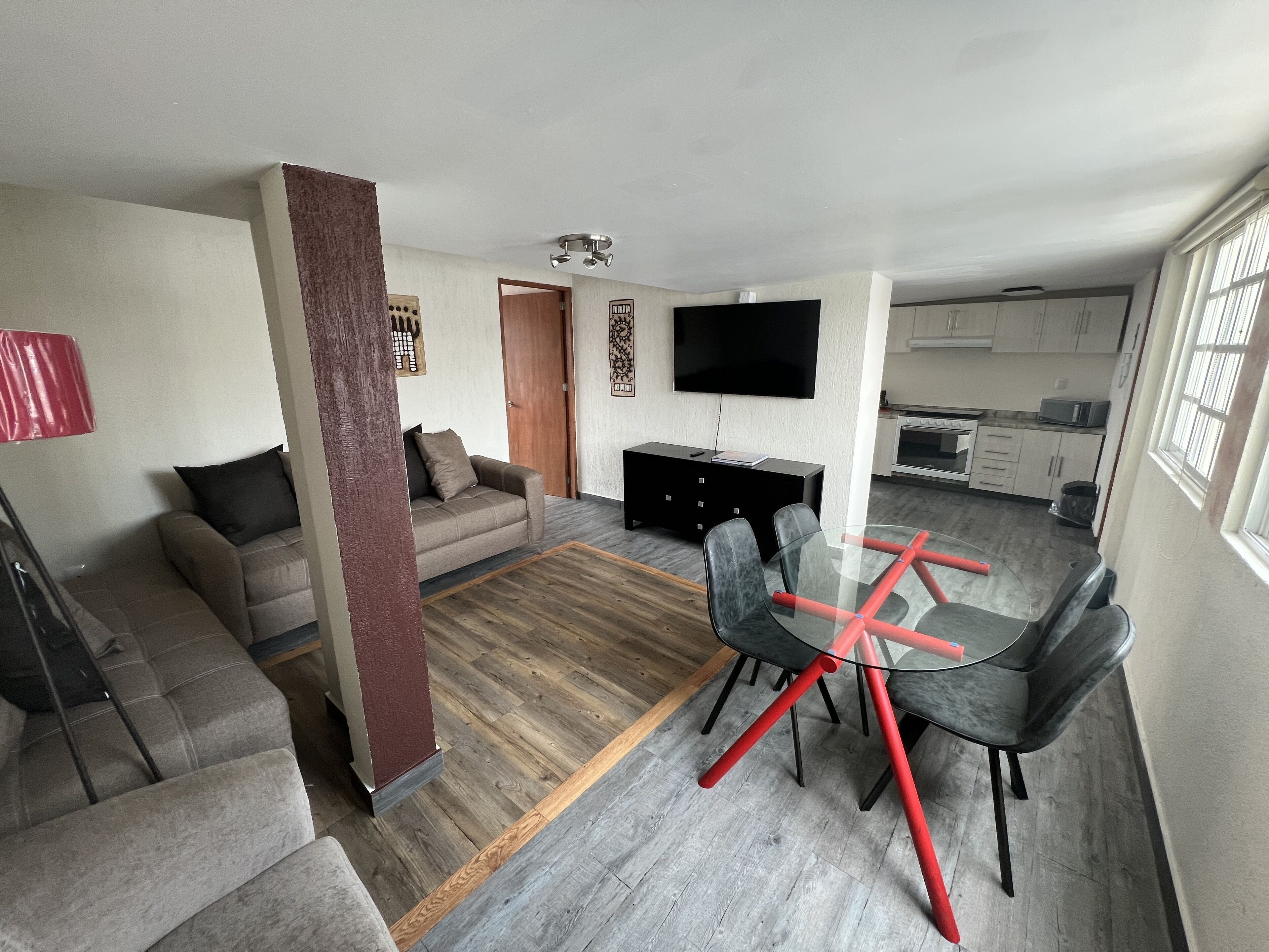 A modern living room with a sofa, a glass dining table with chairs, a TV, and a kitchen area in the background.