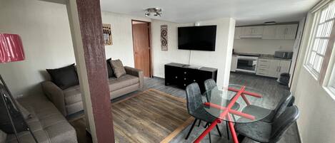 A modern living room with a sofa, a glass dining table with chairs, a TV, and a kitchen area in the background.