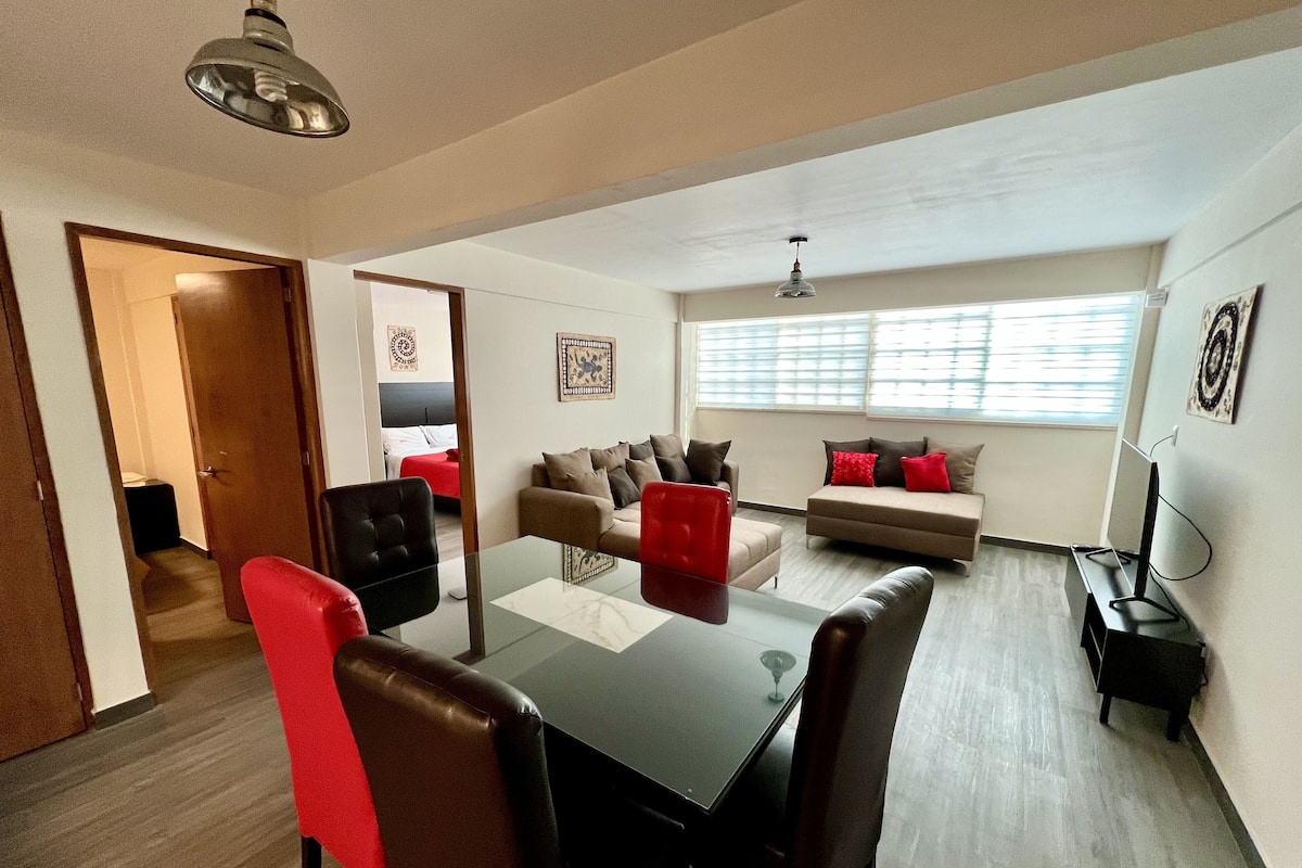 Un salon moderne avec une table à manger en verre, des fauteuils en cuir et un canapé agrémenté de coussins rouges. On y trouve une fenêtre avec des stores, un luminaire au plafond et une porte donnant sur une autre pièce.