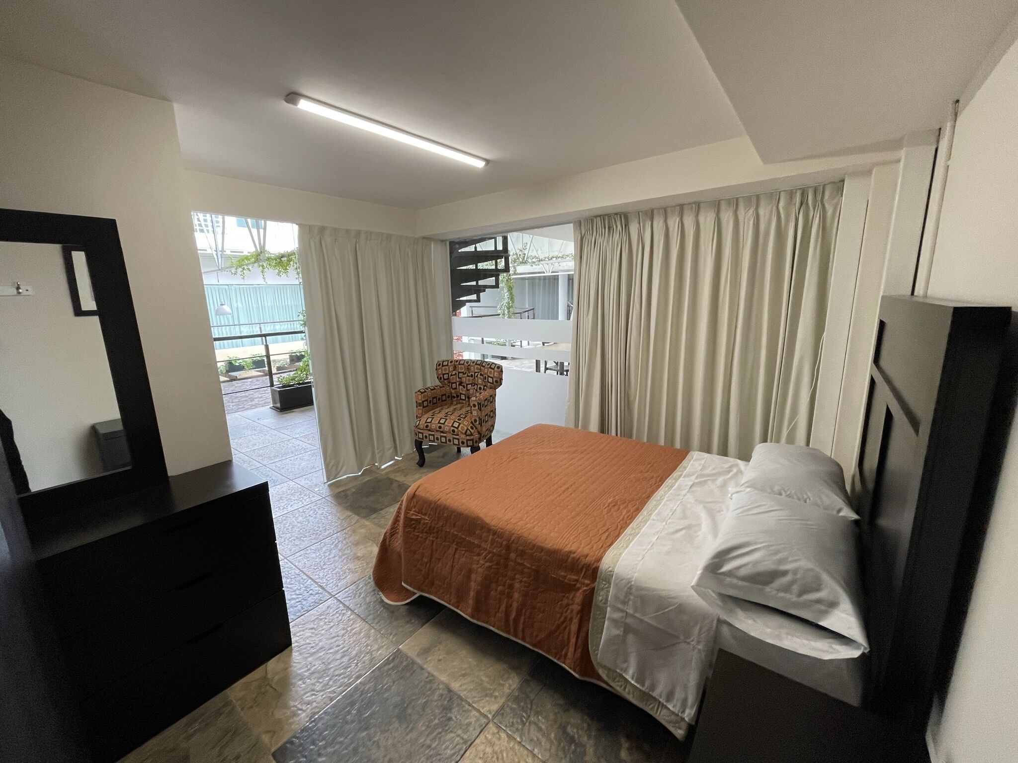 A hotel room with a bed, a chair, a desk, and a balcony with curtains.