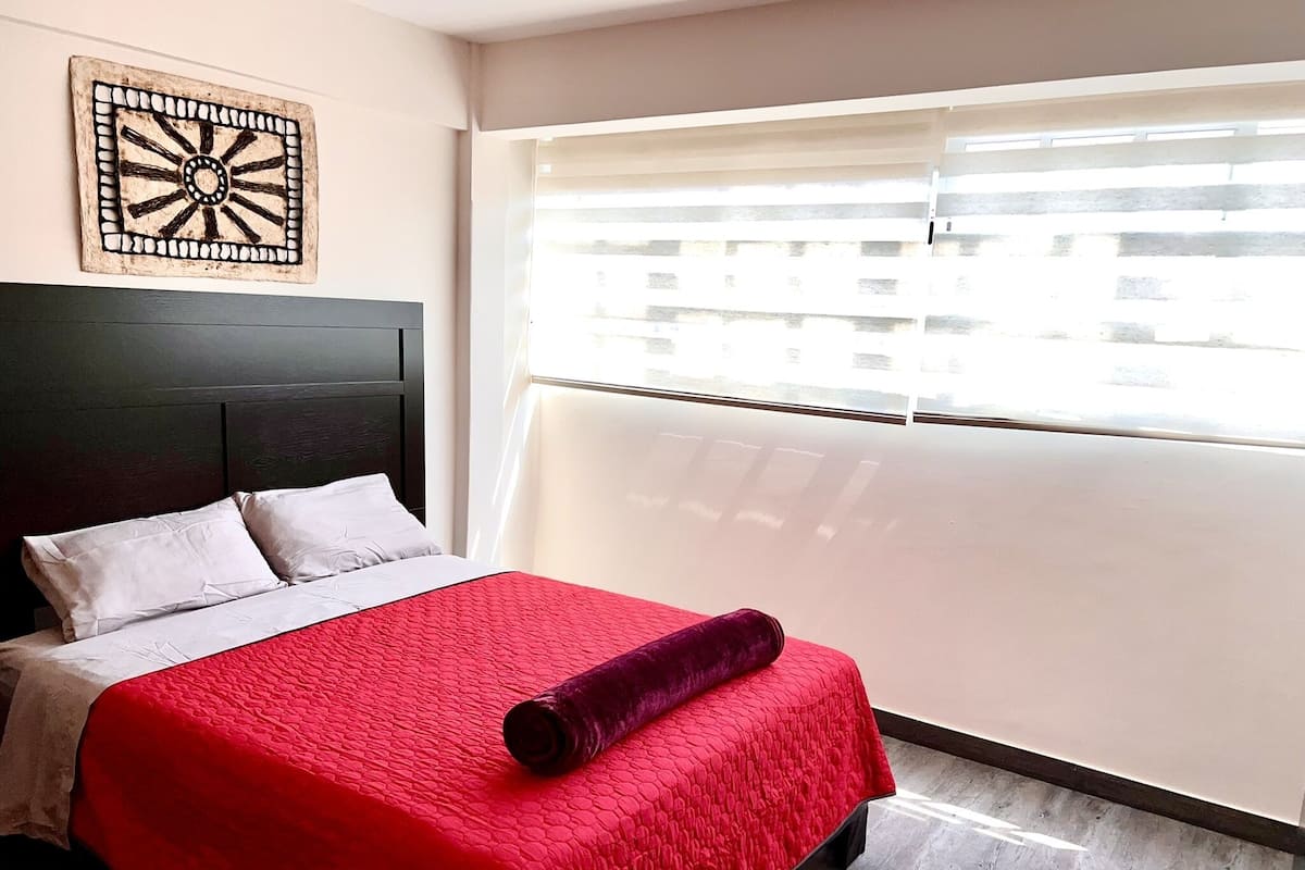 Un lit bien fait, avec une couverture rouge et un plaid violet enroulé, une tête de lit sombre et un élément décoratif fixé au mur.