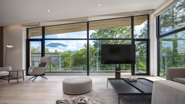 A modern living room with a large TV, comfortable seating, and a view of trees and mountains through large windows. - Ryoun by H2 Life (Kutchan)