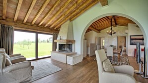 Un salon spacieux doté d’une cheminée, de poutres en bois et d’une grande fenêtre donnant sur un espace verdoyant.