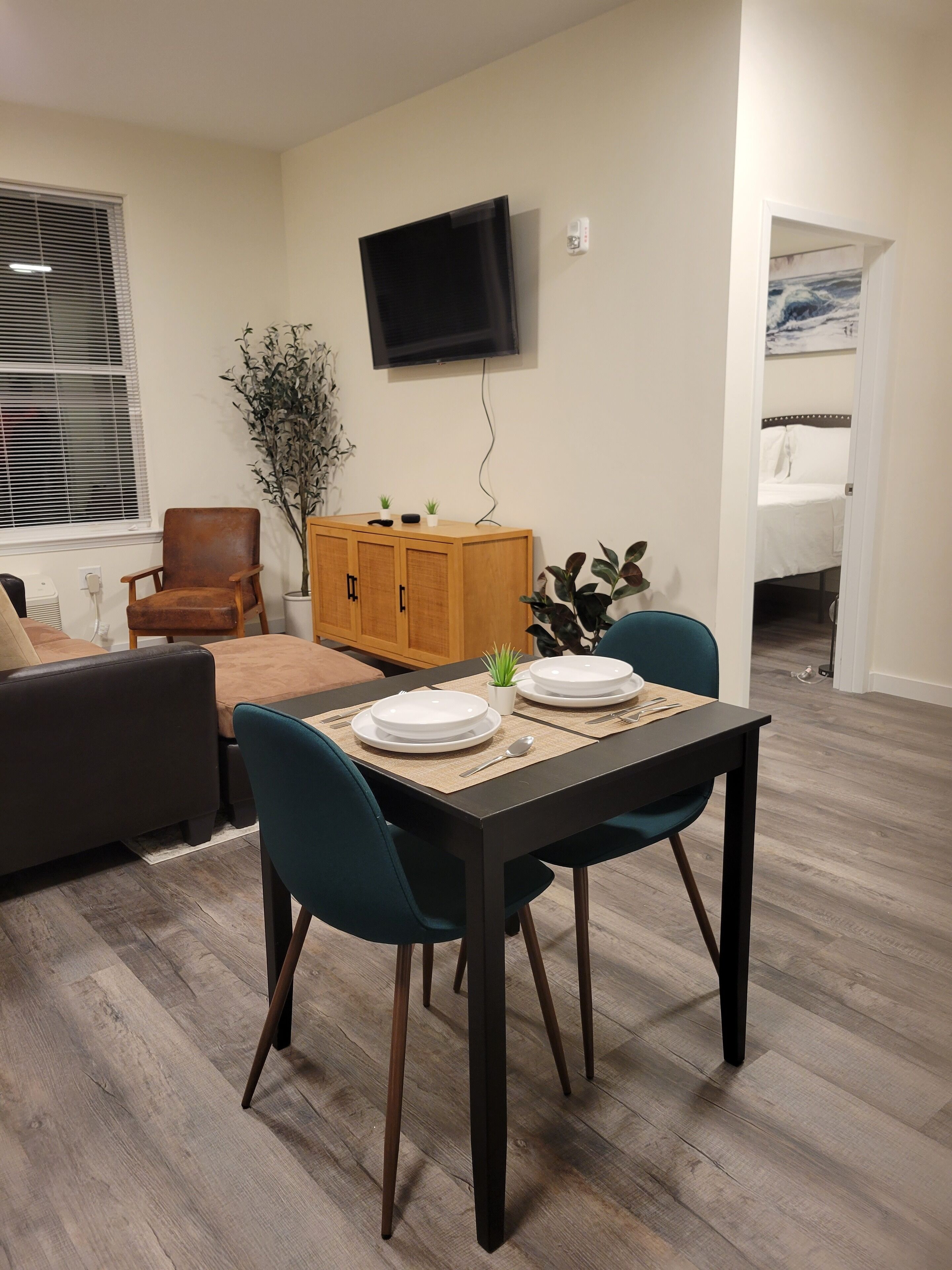 Un coin repas avec une table dressée pour deux personnes, un meuble en bois, un téléviseur fixé au mur et une fenêtre avec des stores.