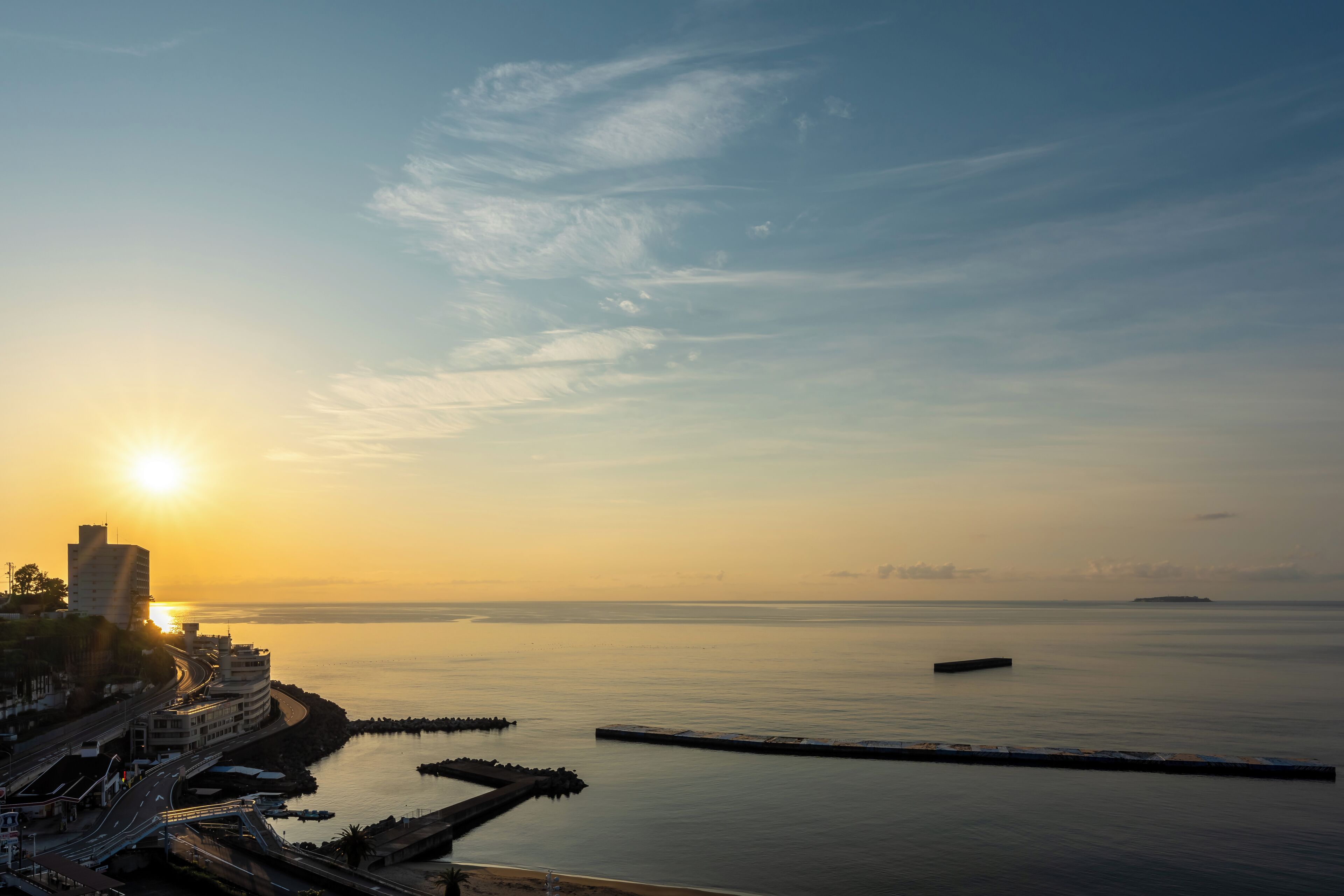 A coastal cityscape at sunset with a calm sea, a pier, and buildings along the shore.