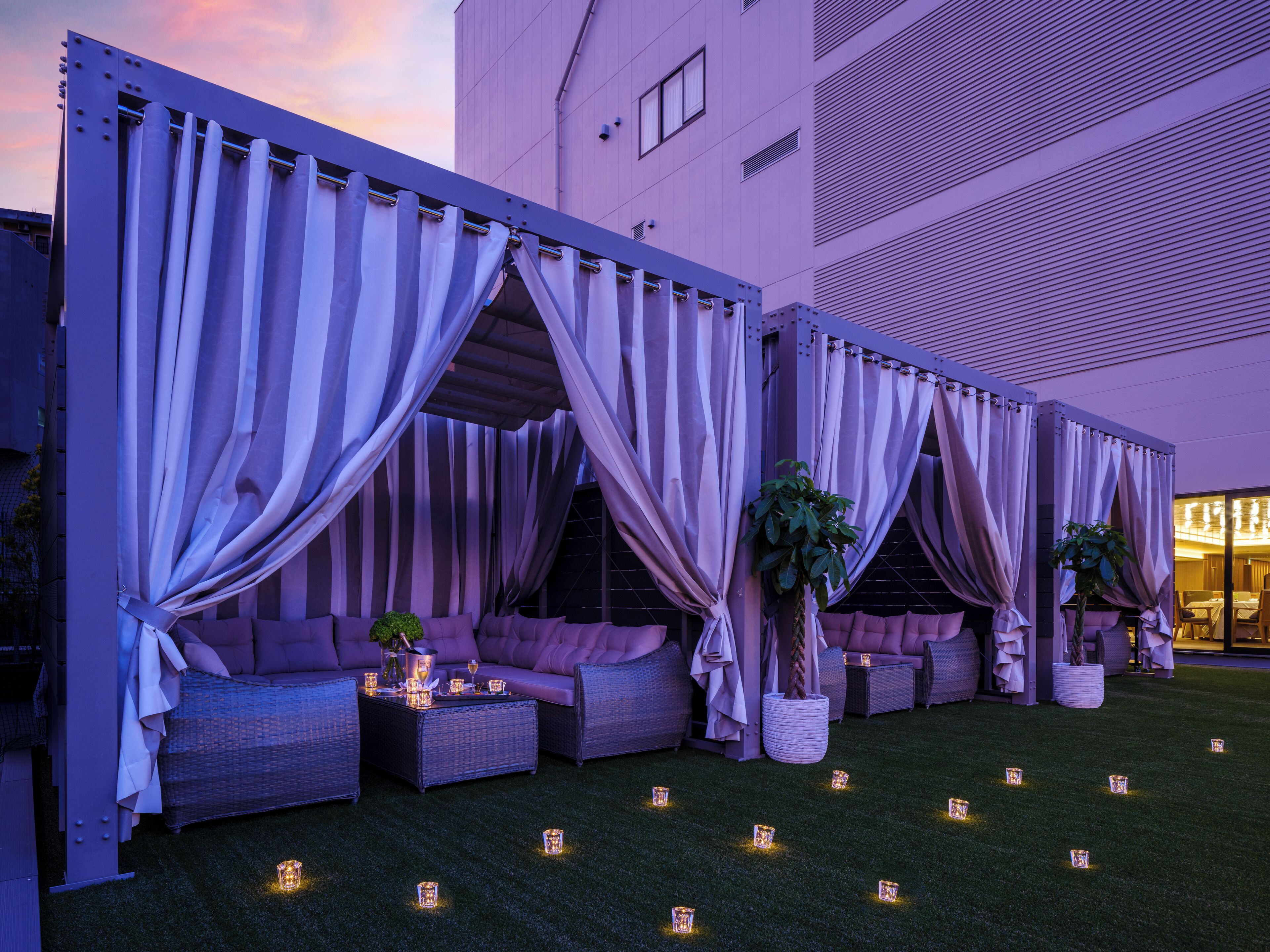 Un espace extérieur aménagé, à moitié abrité, avec des canapés confortables et des tables, entouré de rideaux et décoré de petites guirlandes lumineuses.