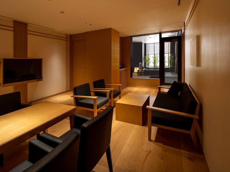 A modern meeting room with a long table, several chairs, a television, and a large window offering a view of the outdoors.