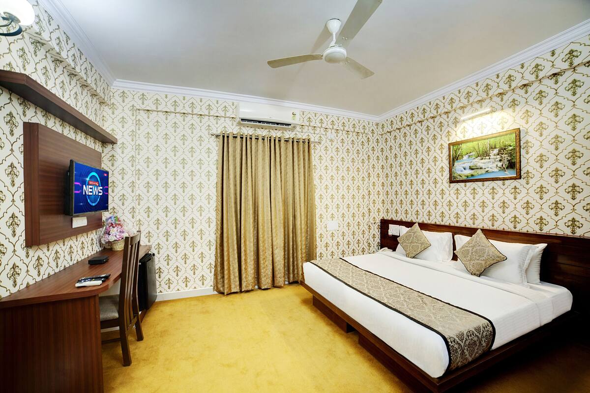 A hotel room with a bed, a desk with a TV, a ceiling fan, and patterned wallpaper.
