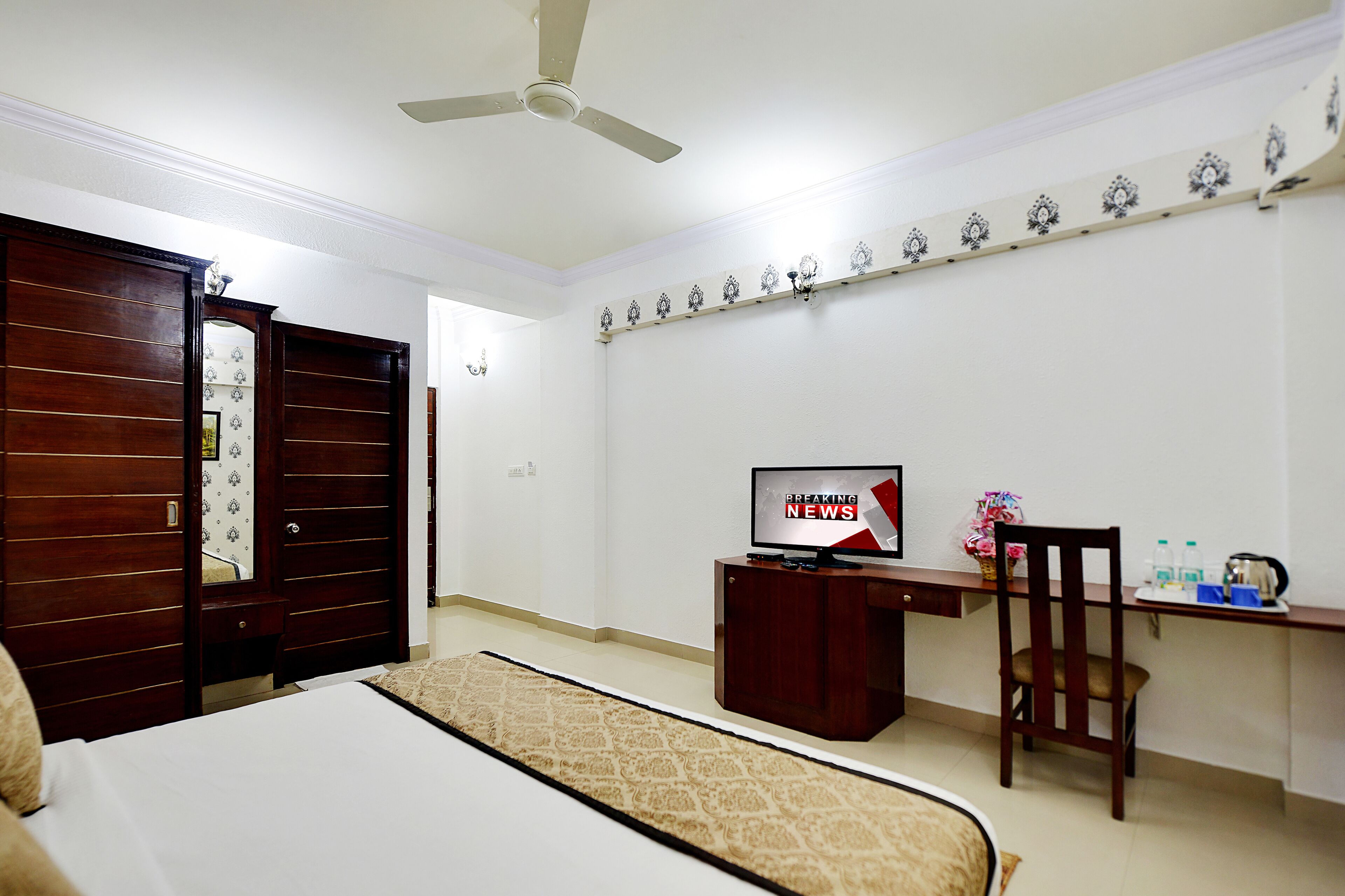 A hotel room with a bed, a desk with a television displaying 'BREAKING NEWS', a chair, and a ceiling fan.