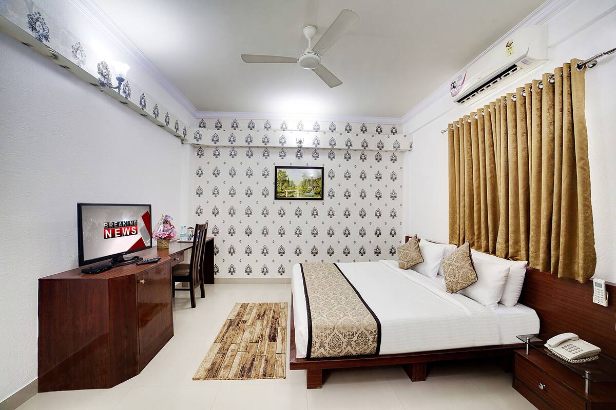 A hotel room with a bed, a desk with a TV, a chair, a rug, and a window with curtains.