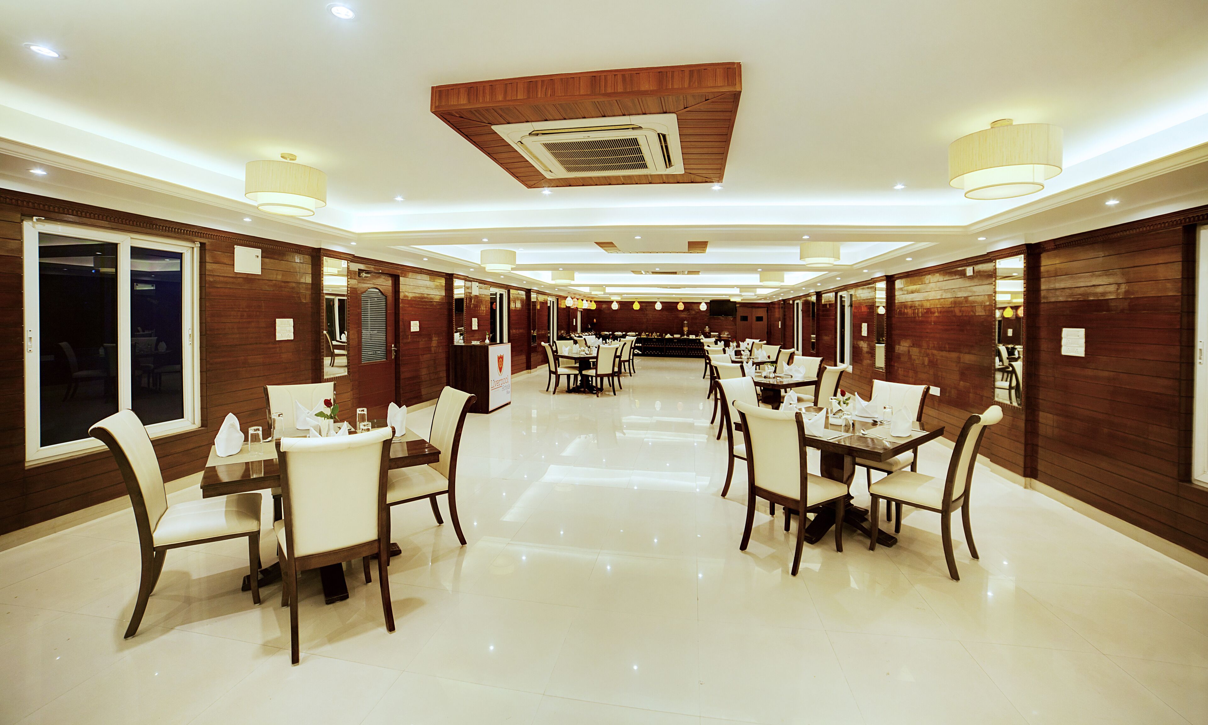 A spacious dining area with multiple tables and chairs, wooden paneling on the walls, and ceiling lights.
