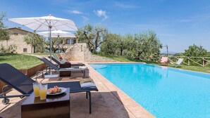 A poolside area with lounge chairs, a table with a plate of food and drinks, and a large umbrella. - Le Residenze dei Potti 12 (Todi)