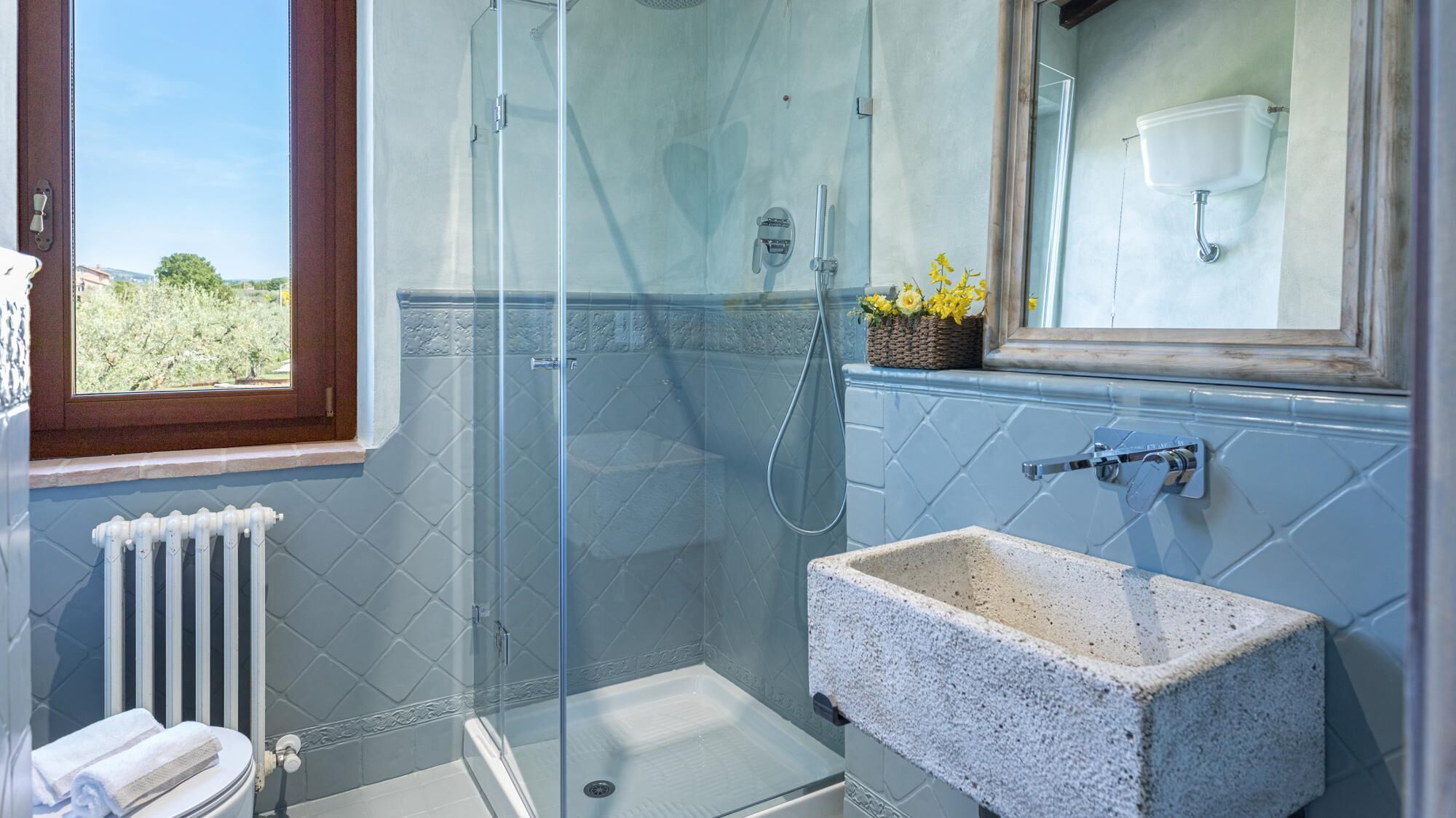 A bathroom with a glass-enclosed shower, a white sink, and a window with a view of greenery.