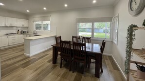 A modern kitchen with white cabinets, a dining area with wooden chairs, and a large window providing a view of the outdoors. - Central Murtoa Home with Lake view, family friendly. (Murtoa)