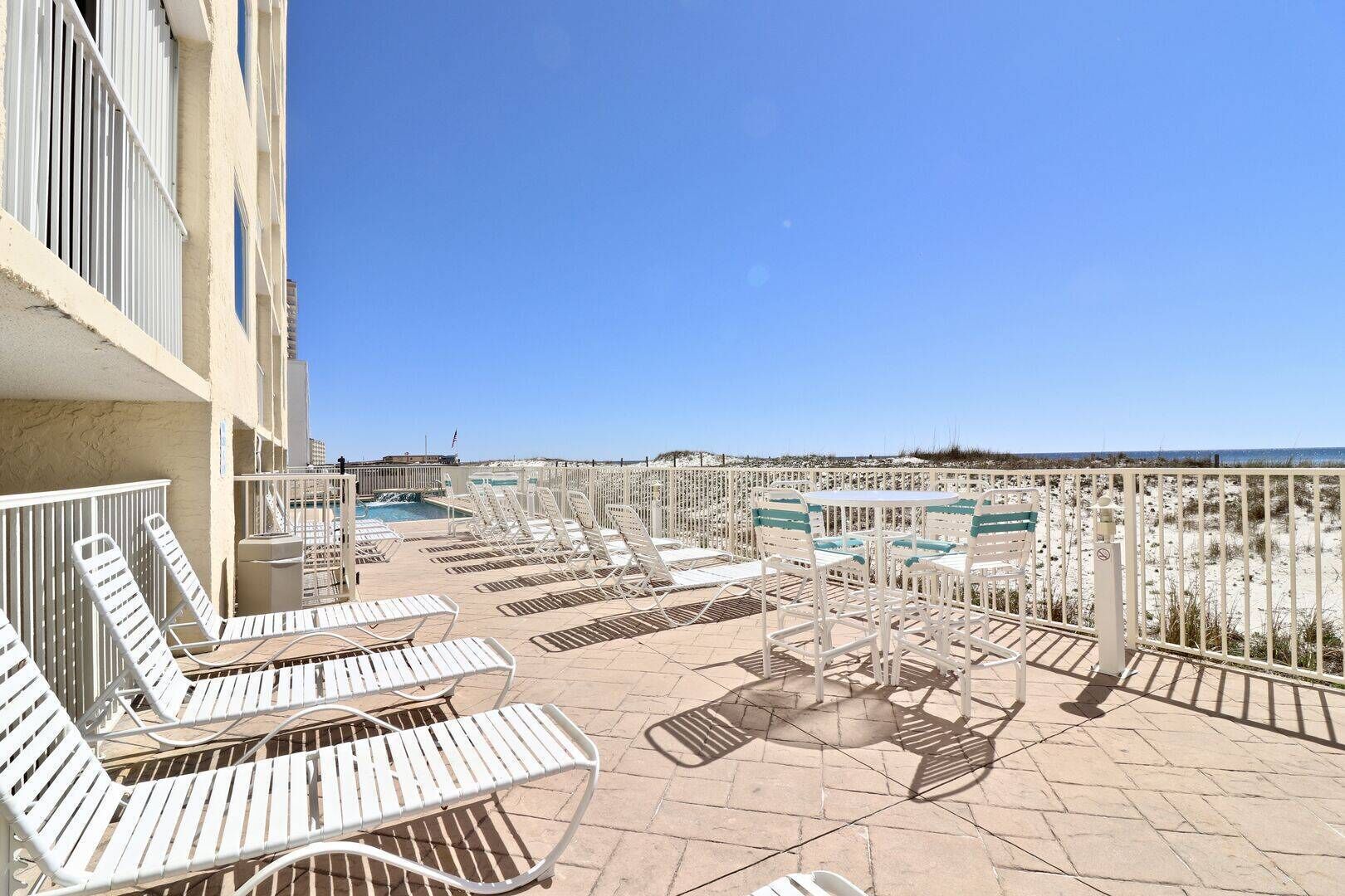A rooftop pool area with white lounge chairs and tables, a clear view of the beach, and a flag on the building.