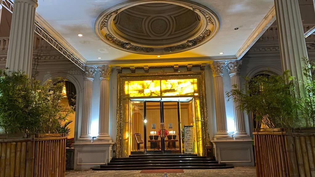 A grand entrance with a large glass door, flanked by columns, leading to a reception area with a chandelier and a signboard.