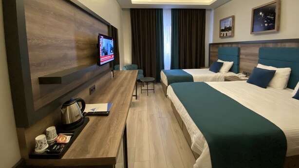 A modern hotel room with two beds, a desk with a coffee maker, a TV mounted on the wall, and a large window with curtains.
