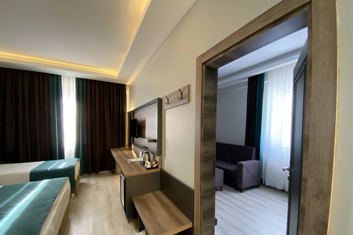 Una habitación de hotel moderna con un escritorio de madera, una cama con cubrecama verde, un área de descanso y vistas a la habitación contigua.