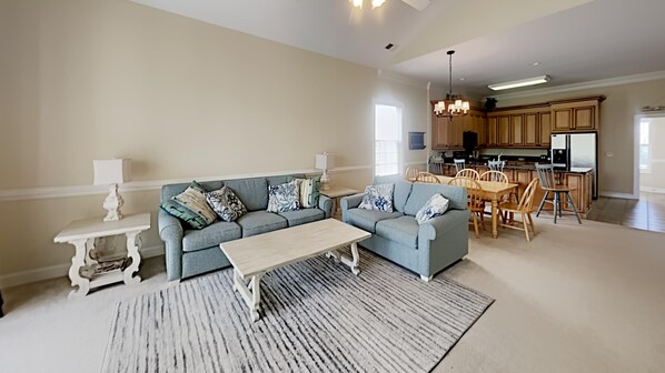 Un salon avec un canapé gris, une table basse et un tapis. On y trouve également un coin repas avec des chaises et une table en bois. La cuisine est visible au fond, avec des meubles en bois et un réfrigérateur.