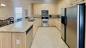 Una cocina con armarios de madera clara, encimera de granito y electrodomésticos de acero inoxidable, incluyendo un refrigerador, horno y microondas.