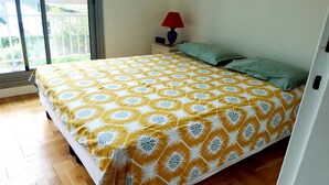 Ein Schlafzimmer mit einem Bett, das mit einer gemusterten Tagesdecke bezogen ist, einem Nachttisch mit Lampe und Radio sowie einem Blick ins Freie durch ein Fenster.