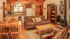 A cozy living room with a sofa, wooden chairs, a coffee table, and a bookshelf. There is a kitchen area with wooden cabinets and a window. - Cute, spacious rainforest cottage close to HVNP (Volcano)