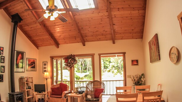 A cozy living room with wooden furniture, a fireplace, and large windows. - Cute, spacious rainforest cottage close to HVNP (Volcano)