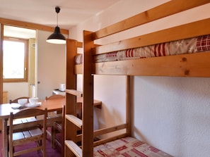 Uma cama beliche com estrutura de madeira e colchão estampado. Há uma mesa de madeira com cadeiras e um lustre pendurado acima.