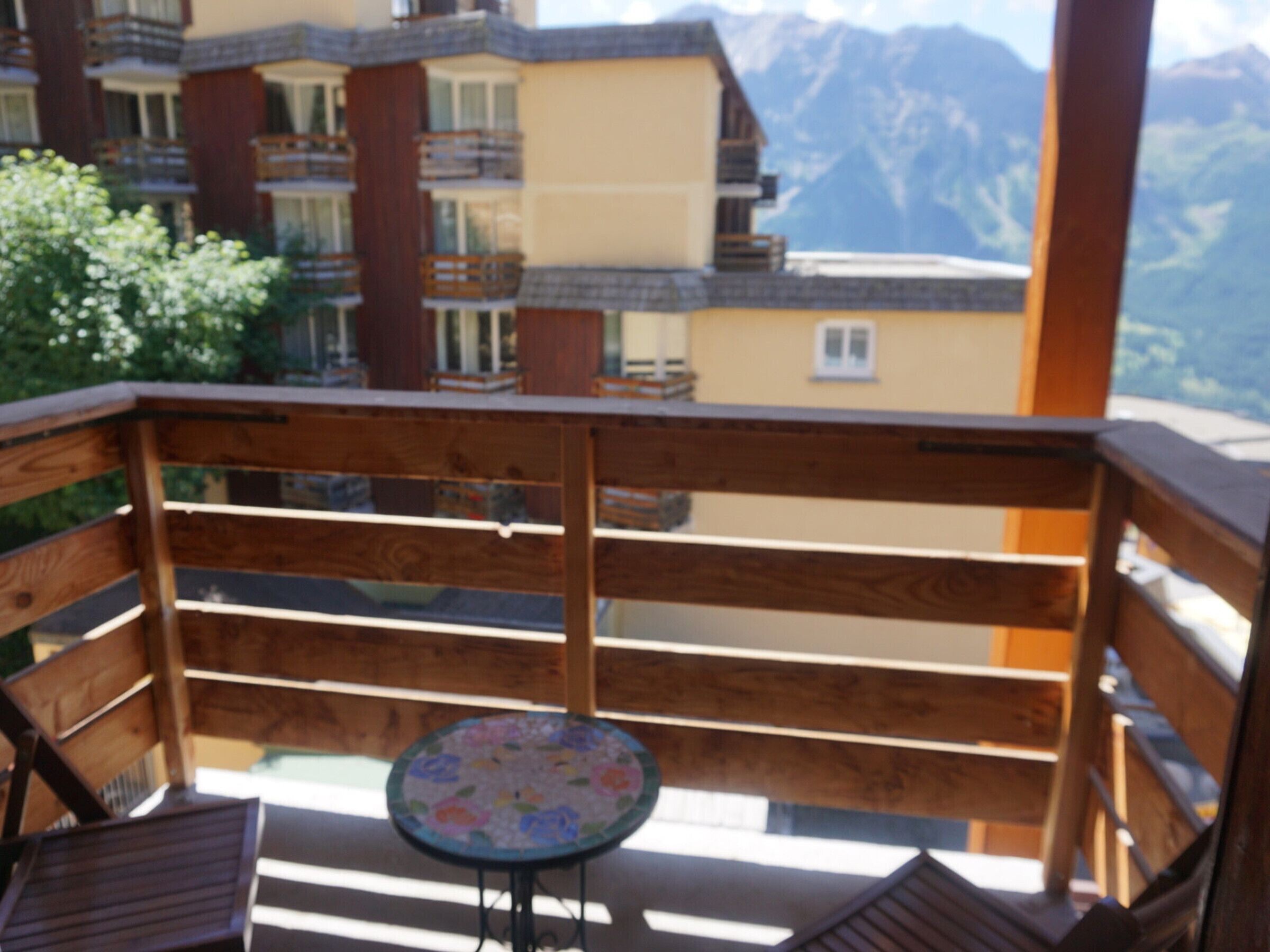 A wooden balcony with a small table and chairs, overlooking a mountain range.