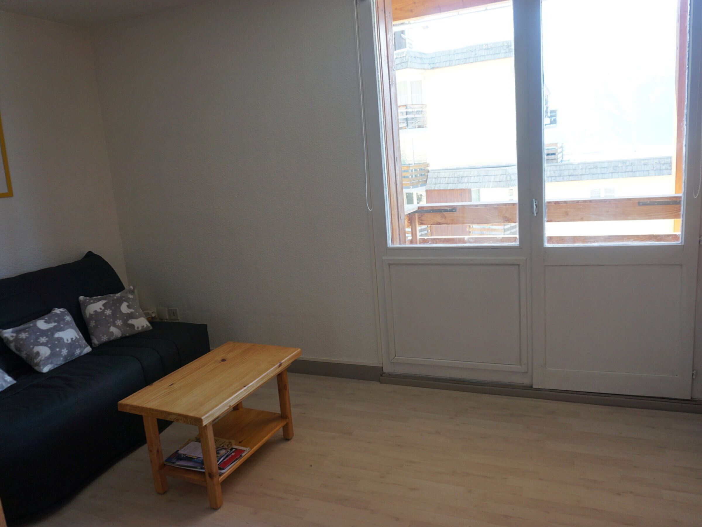 A living room with a dark-colored sofa, two pillows, a wooden coffee table, and a large window with a view of buildings.