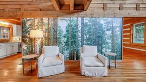 L’intérieur d’un chalet accueillant, avec deux fauteuils blancs, une petite table et une vue sur une forêt, visible à travers de grandes fenêtres.