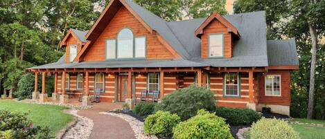 A log cabin with a central chimney, large windows, and a covered porch with seating.