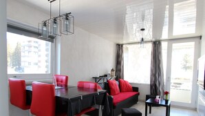 Ein modernes Wohnzimmer mit Essbereich, mit einem schwarzen Esstisch, roten Stühlen, einem schwarzen Sofa und einem schwarzen Hocker.