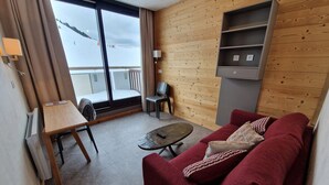 A modern living room with a red sofa, a small table, and a chair. There is a sliding glass door leading to a balcony with a view of a snowy landscape.