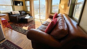 Ein Wohnzimmer mit einer braunen Ledersofa, einem kleinen Tisch und einem roten Hocker. Zwei Fenster mit Vorhängen, ein an der Wand montierter Fernseher und ein gemustertes Teppich sind ebenfalls vorhanden.