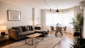 Una sala de estar moderna con un sofá seccional, una mesa de centro, una mesa de comedor con sillas, una lámpara de pie y un ventanal con cortinas.