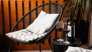 Una silla de mimbre con un cojín, una mesa con una botella y vasos, y una planta en maceta.