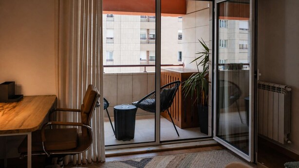 Un apartamento moderno con un puertas corredera de vidrio que da a un balcón, una mesa de comedor de madera, una silla negra y una planta en maceta.