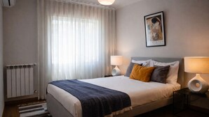 Habitación de hotel con cama, dos lámparas de noche, un radiador, una ventana con cortinas y una obra enmarcada en la pared.