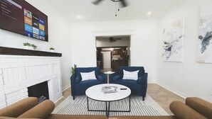 Una sala de estar con chimenea de ladrillo blanco, un sofá azul, una mesa de centro redonda y un televisor de pantalla plana montado en la pared.