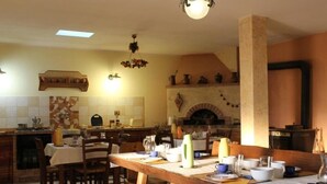 On aperçoit un coin repas avec une longue table en bois, dressée pour quatre personnes, entourée de chaises en bois. Au fond, on distingue une cuisine avec un mur recouvert de carreaux et un four en briques.