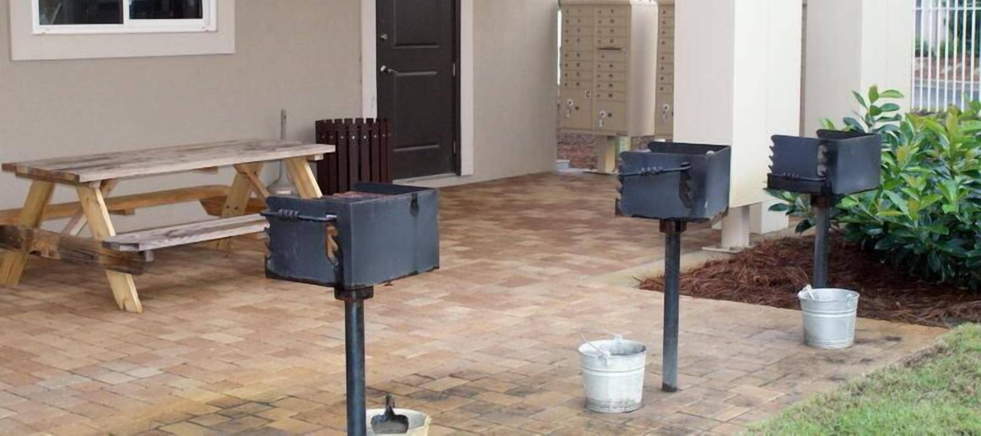 Un espace patio avec un banc en bois, deux supports à boîtes aux lettres et un seau en métal posé sur le sol.