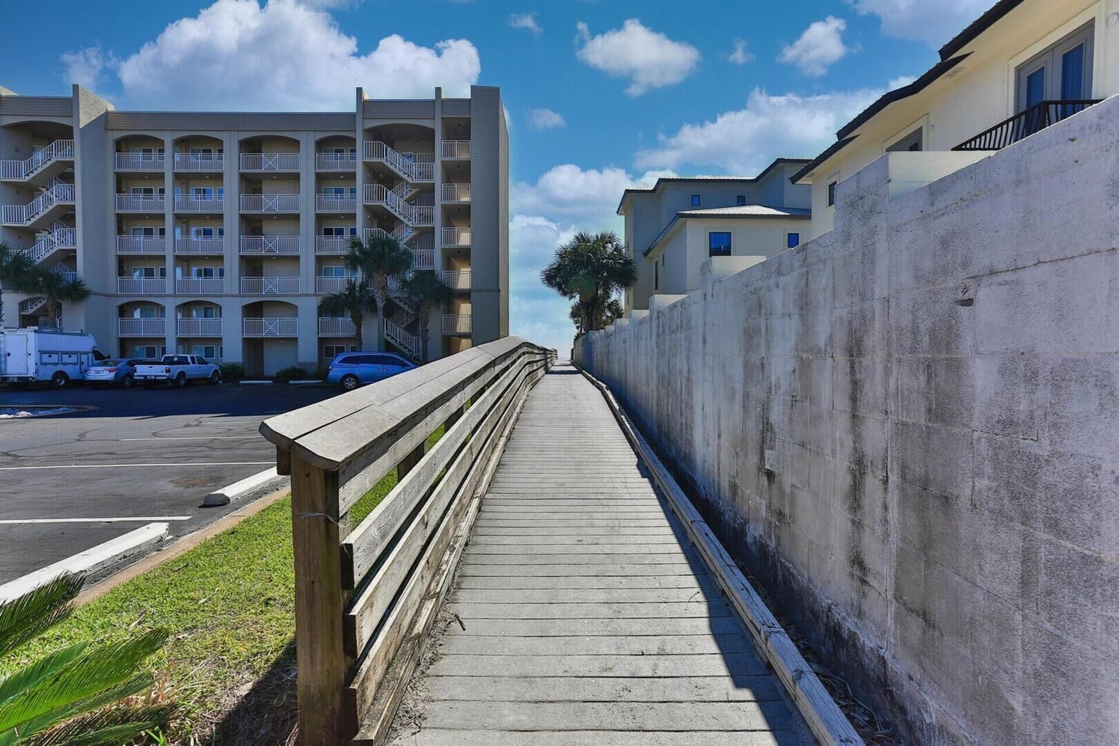 Une promenade en bois mène à un immeuble à plusieurs étages doté de balcons et à un espace de stationnement où se trouvent des véhicules.