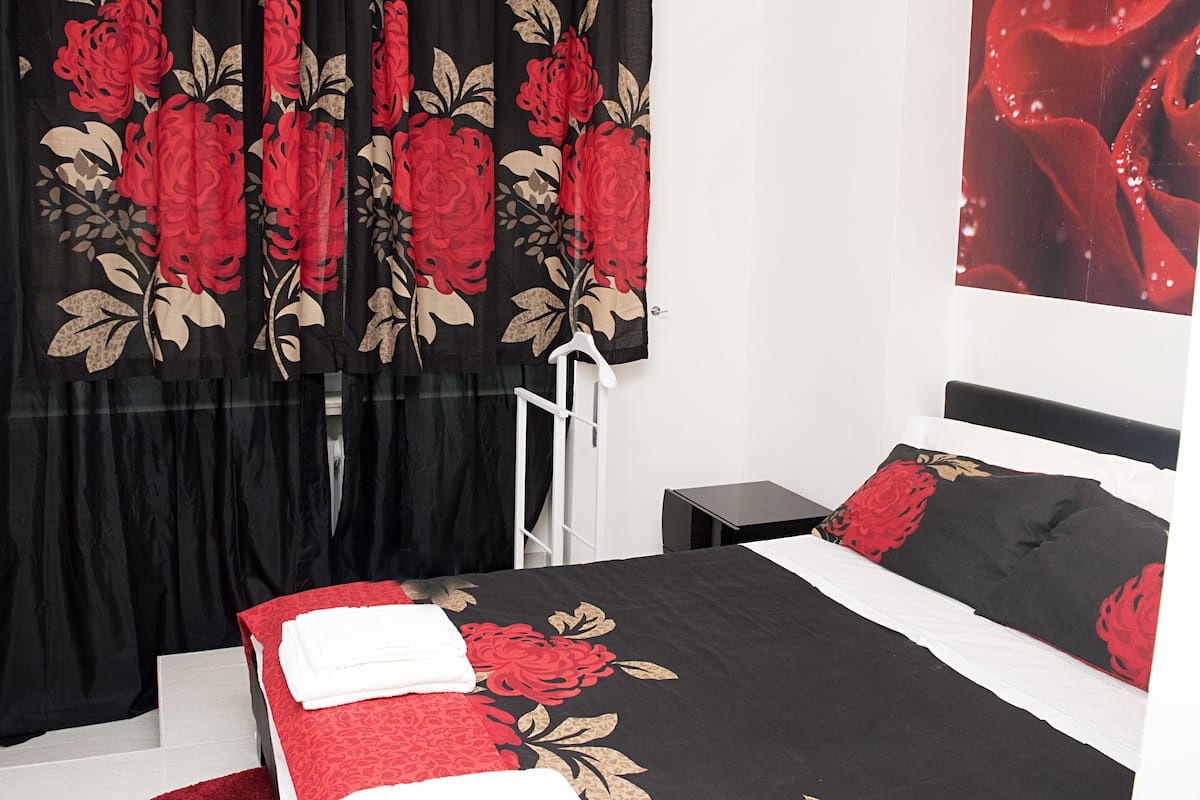 A hotel room with a bed, a nightstand, and a window with black curtains featuring red floral patterns.