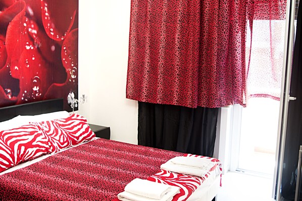 Une chambre d’hôtel avec un lit, une table de chevet, une couvre-lit à motifs rouges et blancs, un rideau rouge et une fenêtre donnant sur un paysage.