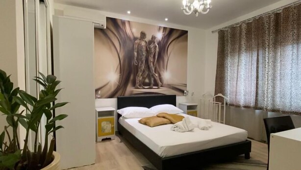 Une chambre moderne avec un lit, une table de chevet, une plante en pot, une fresque murale et une fenêtre avec des rideaux.