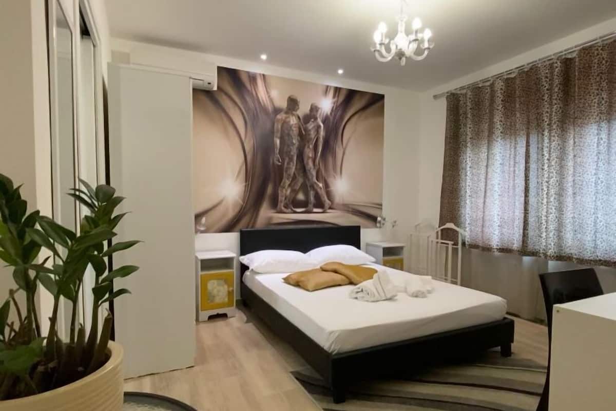 A modern bedroom with a bed, a nightstand, a potted plant, a wall mural, and a window with curtains.